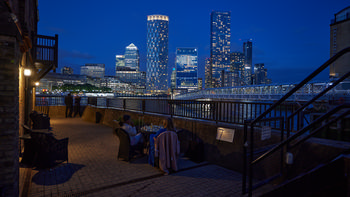 This urban architecture photograph captures an evening scene at a Thames-side restaurant in London during the summer season, with the main subject being the Canary Wharf riverside dining area. The image shows people seated at outdoor tables overlooking the River Thames, while others stand near the railing enjoying the view. Illuminated modern skyscrapers, including One Canada Square and the distinctive cylindrical Newfoundland Tower, dominate the skyline of Canary Wharf in the background. The lights from the buildings are reflected on the river, highlighting the vibrant atmosphere of London’s financial district at dusk. The overall composition emphasizes the blend of contemporary architecture and riverside urban life along the Thames.
